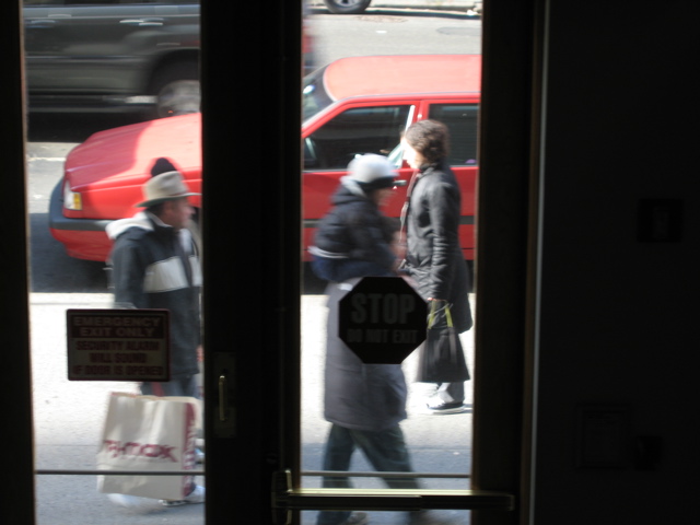 A woman passes a man with a child on his shoulders and a man with a TJ Maxx bag; An SUV passes all