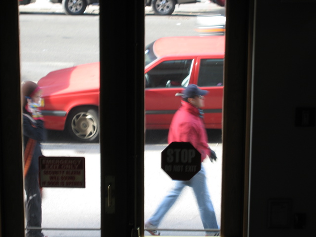 A woman on her cell phone behind a man in a red blazer; A car almost present going south