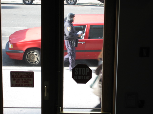 The policeman walks away, white book in hand; a woman walks past in the foreground