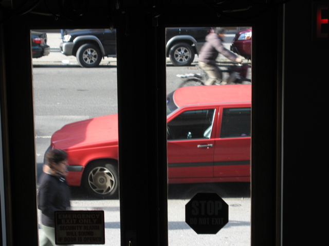 A man walks in the foreground, a man bikes in the background; The SUV in the background begins pulling out of its spot